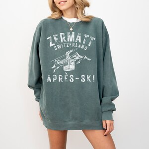 May include: A teal blue sweatshirt with a white graphic print of a ski lift in the mountains. The text "Zermatt Switzerland" and "Apres-Ski" are printed on the sweatshirt.