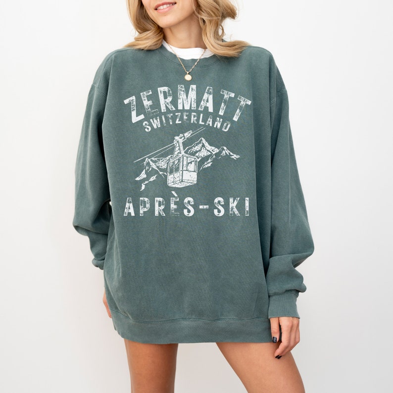 May include: A teal blue sweatshirt with a white graphic print of a ski lift in the mountains. The text "Zermatt Switzerland" and "Apres-Ski" are printed on the sweatshirt.