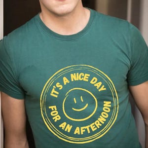 May include: Green t-shirt with a yellow circular graphic. The graphic features a smiling face and the text "IT'S A NICE DAY FOR AN AFTERNOON" in a circular design. The shirt is a solid color and has a crew neck.