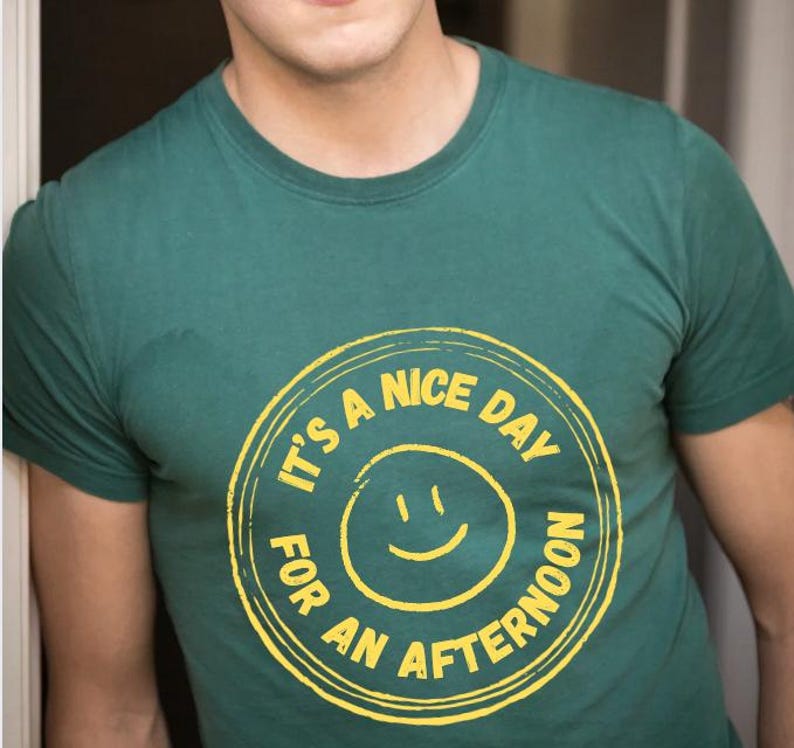 May include: Green t-shirt with a yellow circular graphic. The graphic features a smiling face and the text "IT'S A NICE DAY FOR AN AFTERNOON" in a circular design. The shirt is a solid color and has a crew neck.