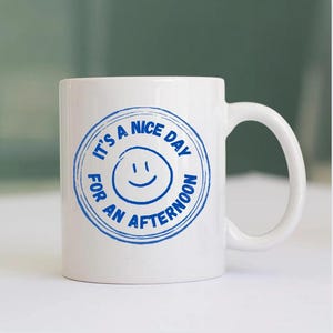 May include: White ceramic mug with a blue circular design that reads "It's a nice day for an afternoon" around a smiling face. The mug has a curved handle and is set against a blurred background.