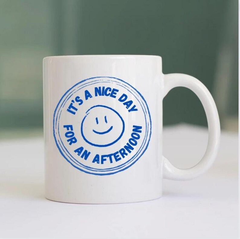 May include: White ceramic mug with a blue circular design that reads "It's a nice day for an afternoon" around a smiling face. The mug has a curved handle and is set against a blurred background.