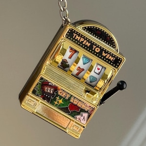 May include: Gold-colored slot machine keychain with the text "TAPIN TO WIN!" and "GET LUCKY!". The slot machine displays sevens, diamonds, and playing card symbols. A black handle is on the side.