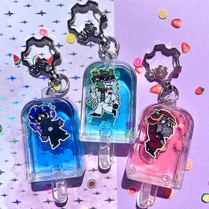 Puede incluir: Tres llaveros transparentes con forma de paleta con diseños de personajes de dibujos animados. Un llavero tiene líquido azul y un personaje con un atuendo negro. Otro tiene líquido azul y un personaje con un traje blanco. El tercero tiene líquido rosa y un personaje con un atuendo rojo.