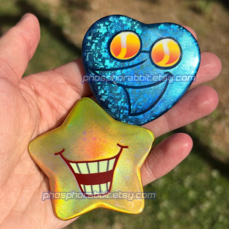 May include: Two holographic stickers: a blue heart with a smiling face and a yellow star with a large grin. The heart has a textured, glittery appearance. The star has a gradient of yellow and green. The text "phosphorabbit.etsy.com" is printed on both.