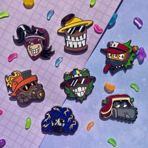 Puede incluir: Una colección de siete coloridos pines de esmalte con diseños de personajes de dibujos animados. Los pines presentan varias caras con gafas de sol, sombreros y expresiones únicas. El fondo es de color púrpura claro con gominolas dispersas.