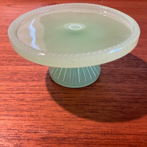 May include: A light green glass cake stand with a round top and a pedestal base. The rim of the top has a decorative beaded edge. The base has vertical ridges. The stand is on a wooden surface.