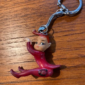 May include: A vintage keychain featuring a small, red elf figurine with a silver-tone metal keyring. The elf has a smiling face, red hair, and is in a seated pose. The keychain is likely made of plastic.