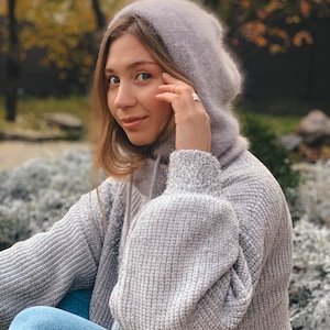 May include: A woman wearing a grey knitted hoodie with a fluffy hood. She is sitting with her legs crossed and looking at the camera.