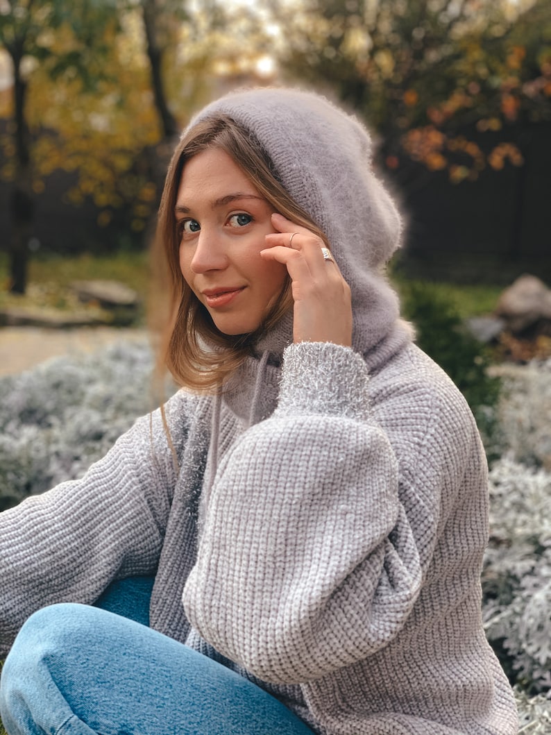 May include: A woman wearing a grey knitted hoodie with a fluffy hood. She is sitting with her legs crossed and looking at the camera.