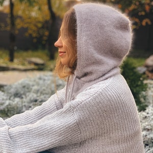 May include: A woman wearing a light purple hooded sweater with a soft, fuzzy texture. The sweater is knitted and has a relaxed fit. The woman is sitting outdoors with a blurred background of trees and greenery.