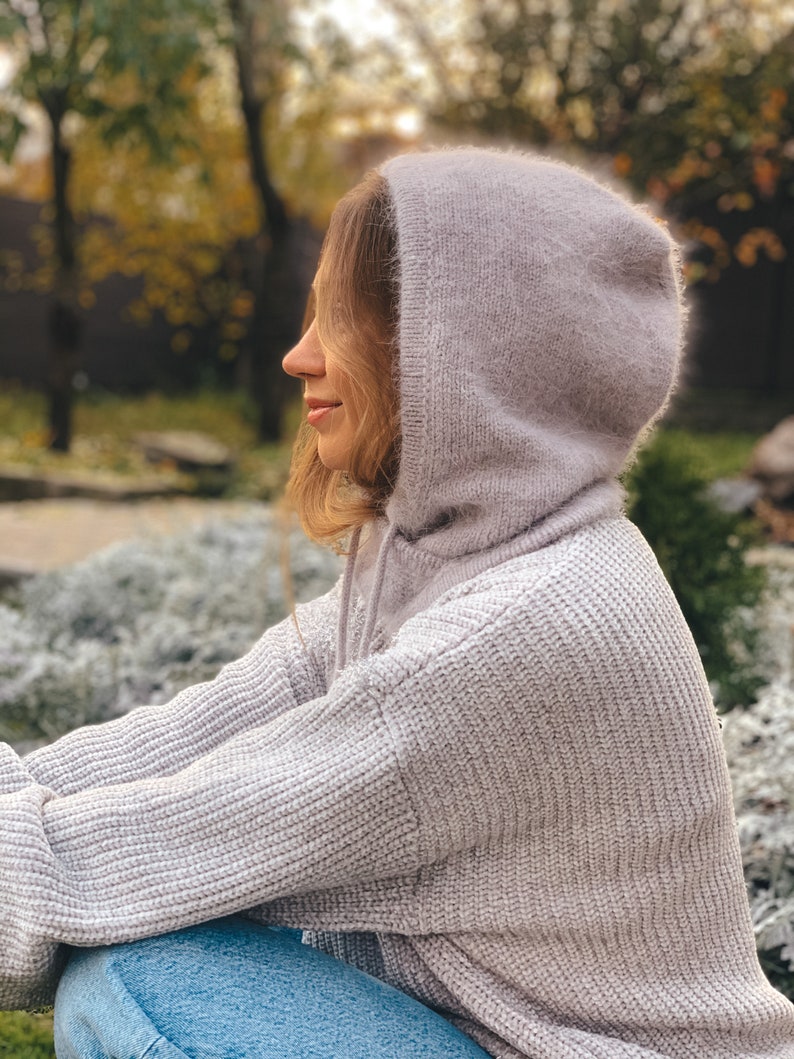 May include: A woman wearing a light purple hooded sweater with a soft, fuzzy texture. The sweater is knitted and has a relaxed fit. The woman is sitting outdoors with a blurred background of trees and greenery.