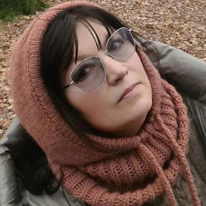 May include: A woman wearing a pink knitted cowl neck scarf and silver-rimmed glasses. The scarf has a cable knit pattern and is tied at the front.