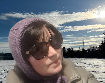 May include: A woman wearing a purple knitted hat and matching scarf with a brown jacket and sunglasses. The background is a snowy landscape with a blue sky and white clouds.
