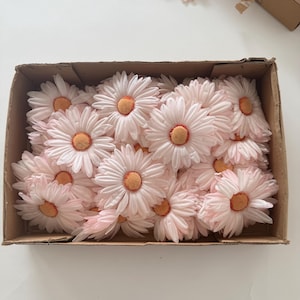 May include: A cardboard box filled with numerous artificial daisy flowers. The flowers have soft pink petals and bright orange centers. The daisies are arranged closely together, filling the box.