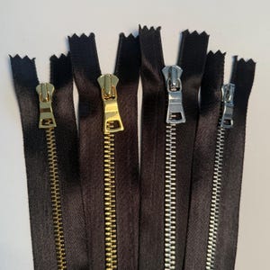 May include: Close-up of several black zippers with various metal zipper pulls. The pulls are in gold and silver tones. The zippers are arranged vertically against a white background.