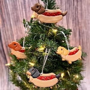 May include: A miniature Christmas tree adorned with four dachshund dog-shaped decorations. The ornaments resemble hot dogs, featuring a bun, a red sausage, and a dachshund dog head and body. The tree is illuminated with small white lights.
