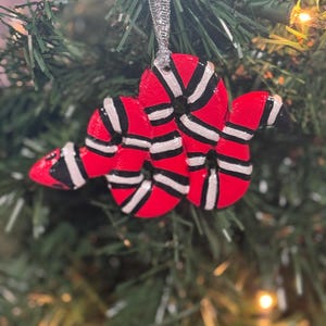 May include: A red, black, and white snake-shaped Christmas ornament. The ornament is hanging from a silver ribbon and is displayed on a green Christmas tree. The snake has a red body with black and white stripes.