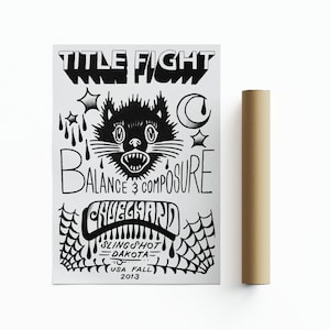 May include: A monochrome poster featuring the words "TITLE FIGHT" and "BALANCE & COMPOSURE" in a distinctive font. The design includes a black cat illustration and other graphic elements. A brown cardboard tube is positioned beside the poster.