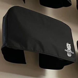 Könnte beinhalten: Schwarze Abdeckung mit dem weißen Text "PINEAPPLE GRIPS". Die Abdeckung hat eine rechteckige Form mit zwei Aussparungen unten. Sie dient wahrscheinlich zum Schutz.