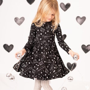Puede incluir: Una niña pequeña con un vestido negro con un estampado blanco de guitarras y las palabras "Love Rocks" y "Live Rocks". Está sosteniendo una cadena de luces de discoteca plateadas.