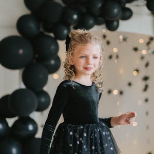 Child smiling in a black star-tulle tutu bodysuit, standing beneath black balloons.