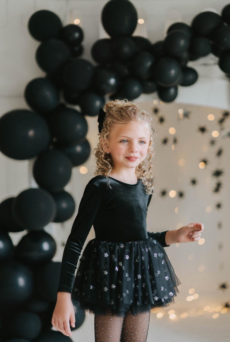 Child smiling in a black star-tulle tutu bodysuit, standing beneath black balloons.