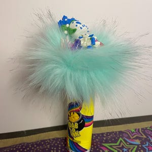 Pu&ograve; includere: Una composizione stravagante con un cilindro giallo con la scritta "Dab O Ink" e un design arcobaleno. &Egrave; sormontato da una soffice pelliccia sintetica azzurra e piccoli oggetti decorativi.