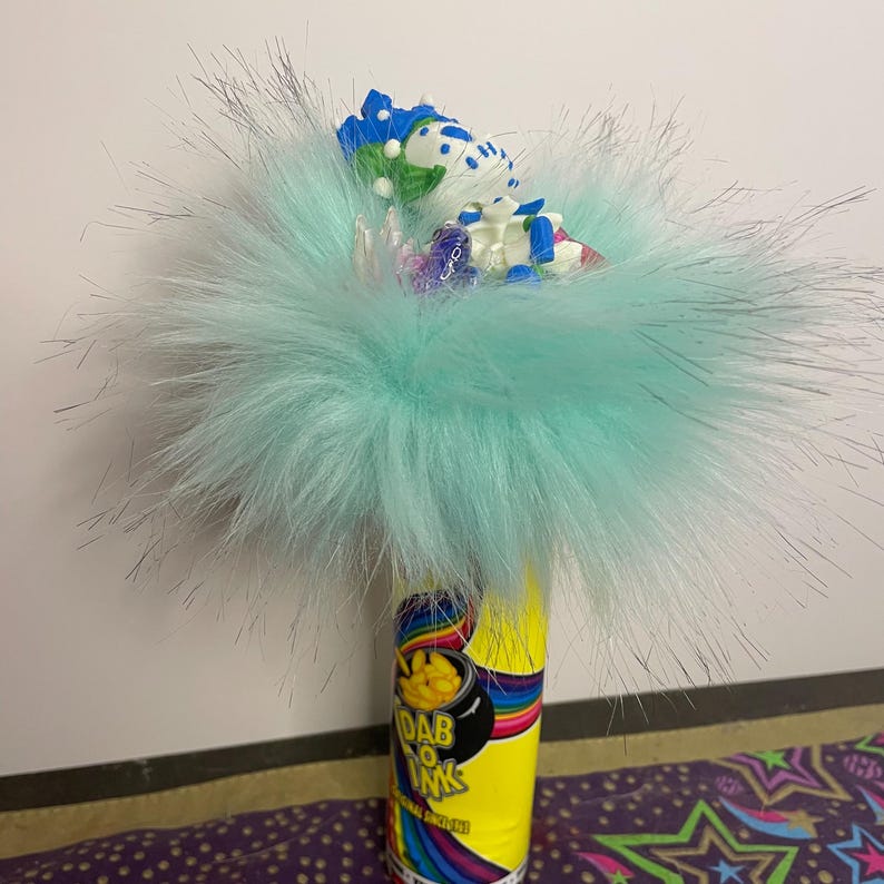 Pu&ograve; includere: Una composizione stravagante con un cilindro giallo con la scritta "Dab O Ink" e un design arcobaleno. &Egrave; sormontato da una soffice pelliccia sintetica azzurra e piccoli oggetti decorativi.