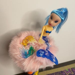 May include: A mermaid doll with blue hair and a blue bikini top sits atop a pink fluffy base. Two plastic fish, one gold and one green, are attached to the base. The mermaid has a blue tail.