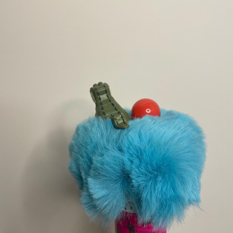 Pu&ograve; includere: Un accessorio stravagante e colorato con un elemento di pelliccia blu chiaro e soffice. Una piccola sfera rossa e un pezzo verde strutturato sono attaccati alla pelliccia. La base dell'articolo &egrave; rosa vibrante con un motivo floreale.