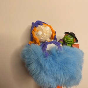 Puede incluir: Una figura caprichosa con tem&aacute;tica de Halloween que presenta una mu&ntilde;eca con cabello naranja, una cara cosida y una falda de piel azul. Una bruja verde con un sombrero negro y una escoba est&aacute; al lado de la mu&ntilde;eca. Fondo liso.