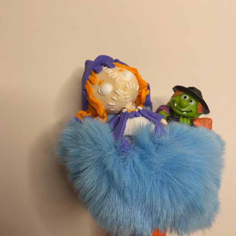 Puede incluir: Una figura caprichosa con tem&aacute;tica de Halloween que presenta una mu&ntilde;eca con cabello naranja, una cara cosida y una falda de piel azul. Una bruja verde con un sombrero negro y una escoba est&aacute; al lado de la mu&ntilde;eca. Fondo liso.