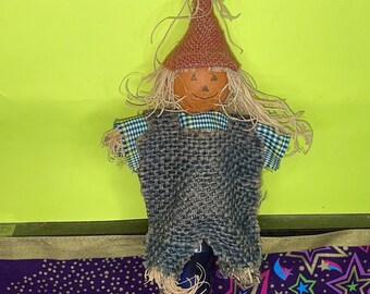 May include: A handmade scarecrow doll with a burlap body, a plaid shirt, and a brown hat with a painted pumpkin face. The scarecrow is standing on a purple and gold fabric with stars.