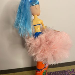 May include: A doll with blue hair and a pink fluffy skirt. The doll is wearing a blue top and bottom. The doll is attached to an orange and red object with a colorful design.