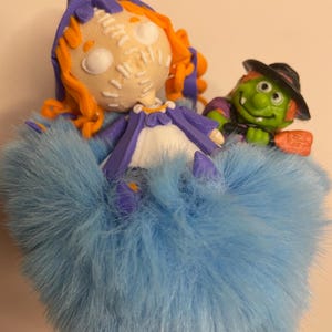 Puede incluir: Una figura caprichosa con tem&aacute;tica de Halloween que presenta una mu&ntilde;eca con cabello naranja, una cara cosida y una falda de piel azul. Tambi&eacute;n hay una bruja verde con un sombrero negro y una escoba. La figura es un art&iacute;culo decorativo &uacute;nico.