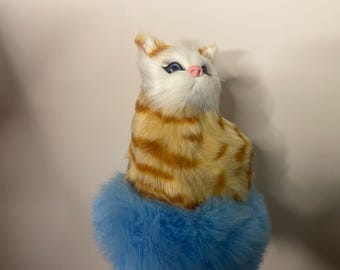 May include: A plush cat figurine with a white face, blue eyes, and a pink nose. The cat has orange and yellow striped fur and sits atop a fluffy blue base. The figurine is a decorative item.