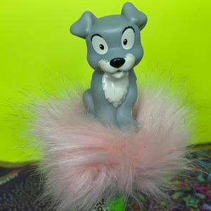 Könnte beinhalten: Eine graue Welpenfigur mit großen Augen und weißer Brust sitzt auf einem flauschigen rosa Pompon. Der Welpe hat schwarze Augen und eine schwarze Nase. Der Hintergrund ist ein leuchtendes Neongrün.