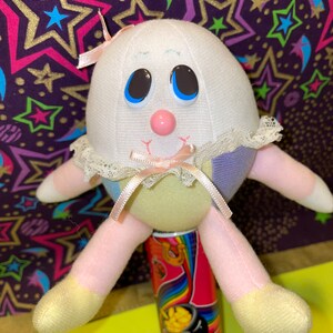 May include: A plush toy shaped like an egg with a pink nose, blue eyes, and a pink bow. The toy is sitting on a red and rainbow can with the text "Dab o Ink" on it.