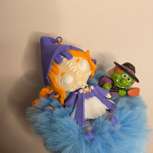 Puede incluir: Un conjunto de figuras caprichosas con tem&aacute;tica de Halloween. La figura principal es una bruja con aspecto de mu&ntilde;eca con cabello naranja, un sombrero y abrigo morados y una cara cosida. Tambi&eacute;n hay una bruja verde con un sombrero negro y una escoba, todo anidado en piel azul.