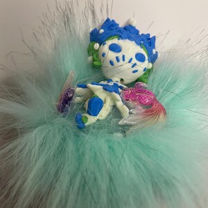Pu&ograve; includere: Un accessorio stravagante fatto a mano con una figura di creatura blu, bianca e rosa su una base soffice azzurra. La figura presenta dettagli intricati ed &egrave; circondata da materiale morbido e piumato, creando un design unico e giocoso.