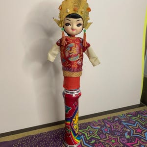 May include: A vintage doll with a traditional Asian-inspired outfit. The doll has a gold headdress, a red top with a dragon design, and a cylindrical base. The base is a red can with colorful graphics.