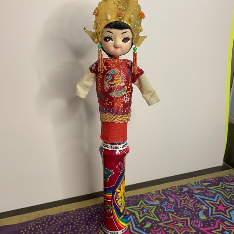 May include: A vintage doll with a traditional Asian-inspired outfit. The doll has a gold headdress, a red top with a dragon design, and a cylindrical base. The base is a red can with colorful graphics.