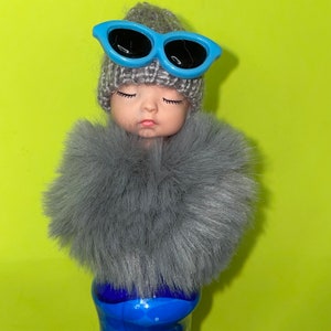 Può includere: Una figura simile a una bambola con gli occhi chiusi, indossa un cappello di lana grigio e occhiali da sole azzurri. Una soffice sciarpa grigia circonda il collo. La figura poggia su una bottiglia di vetro blu e trasparente, su uno sfondo verde brillante.