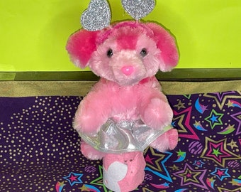 May include: Pink plush toy dog wearing a silver sparkly skirt and silver heart-shaped headbands. The dog is sitting on a purple fabric with a star pattern.