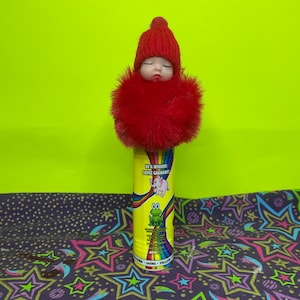 May include: A whimsical doll with a red knitted cap and a fluffy red collar rests atop a yellow cylinder. The cylinder features a rainbow design, an elephant and frog graphic, and the text "BE A WINNER! SOYEZ GAGNANT!"