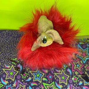 May include: A whimsical dog figurine with a red fur mane. The dog has tan fur, black eyes, and a black nose. The background is a bright green and a patterned fabric with stars.