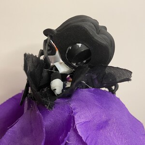 May include: A black, 3D-printed decorative head with a large eye and a ruffled black fabric. The head is attached to a purple fabric base. The design is reminiscent of a Halloween decoration.