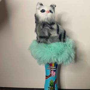 Può includere: Una statuetta di gatto di peluche con strisce grigie e nere è seduta sopra un cilindro avvolto in un materiale soffice verde chiaro. Il cilindro presenta illustrazioni colorate e il testo "BE A WINNER / SOYEZ GAGNANT".