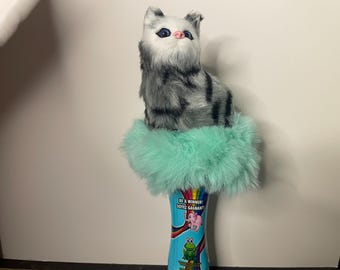 May include: A plush cat figurine with gray and black stripes sits atop a cylinder wrapped in a fluffy, light green material. The cylinder has colorful illustrations and the text "BE A WINNER / SOYEZ GAGNANT".